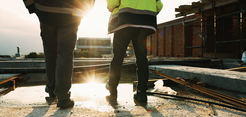 To mænd i arbejdstøj og refleksjakker går på en byggeplads i modlys fra solen.